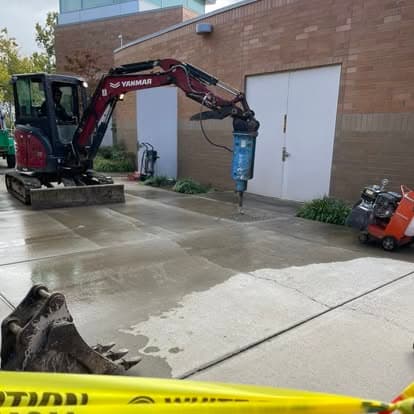 Excavator with hydraulic breaker on concrete site near construction equipment and caution tape.