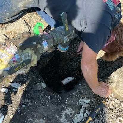 Worker inspecting a buried utility valve in a gravel area, using tools for maintenance.