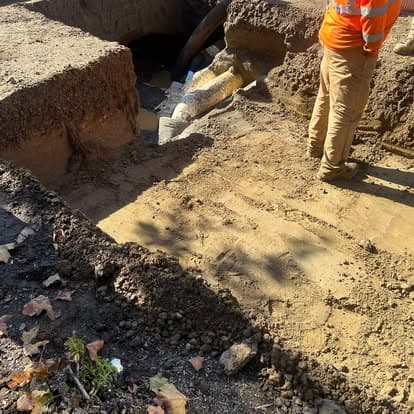 Construction worker inspecting a large excavation site with exposed pipes and surrounding dirt.