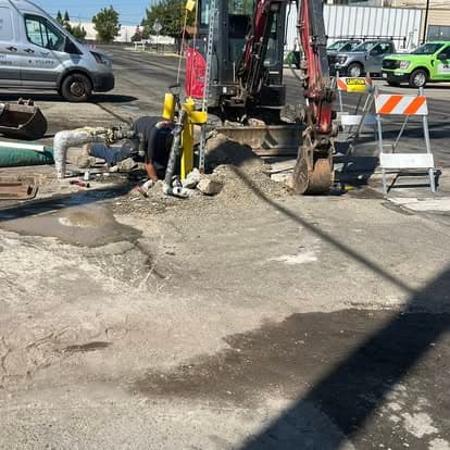 Construction work with heavy machinery on a street, worker digging near a utility line.