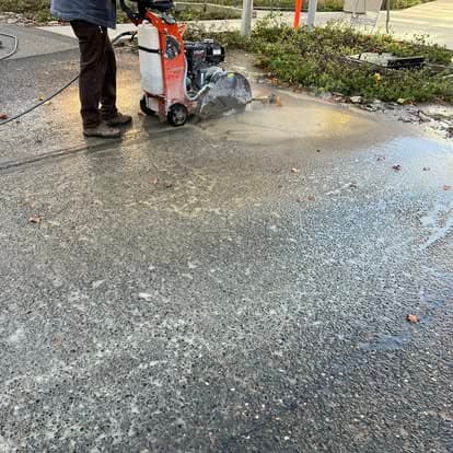 Construction worker using a concrete saw on pavement, with water spraying to control dust.