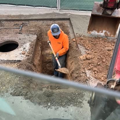 Worker in orange shirt digging in construction site, using shovel near excavation equipment.