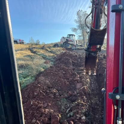 Excavator digging trench in rural landscape with machinery and equipment visible.