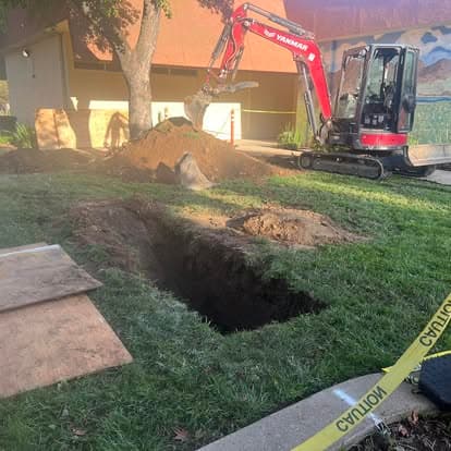 Excavator digging a trench in front of a building, with safety tape visible on the ground.