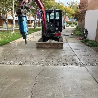 Excavator breaking concrete on a walkway, surrounded by trees and buildings.