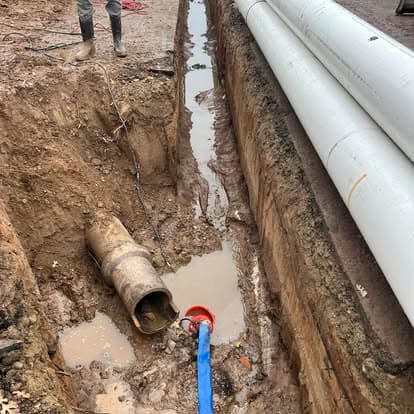 Excavation trench with water, exposed pipe, and construction worker in gray boots.