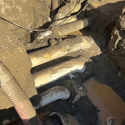 Exposed underground utility pipes in muddy excavation site with water pooling nearby.
